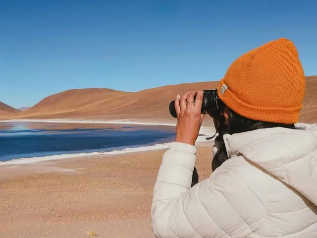 Woman looking through binoculars at a vast desert landscape and lake.